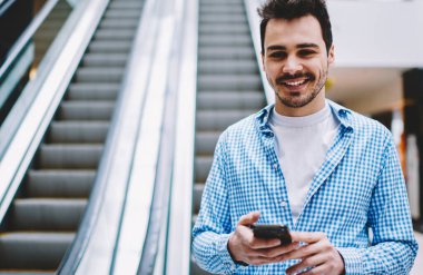 Elinde modern akıllı telefon gadget ile elektrikli merdivenlerde ayakta ve boş zamanlarında iyi hissederken kamera bakarak şık gömlek mutlu gülümseyen hipster adam yarım uzunlukta portre