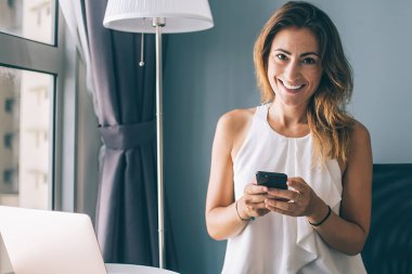 Beyaz kolsuz bluz giyen, akıllı telefondan mesaj atan evde dizüstü bilgisayar kullanan ve kameraya bakan hoş, esmer bir kadın.