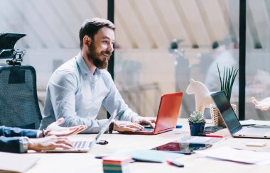 Gülümseyen erkek ve kadın çalışanlardan oluşan bir ekip modern ofiste dizüstü bilgisayarlarla proje üzerinde tartışırken...