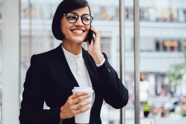 Gözlük neşeli kadın tüccar lezzetli kahve ile açık havada zaman tadını çıkarırken akıllı telefon konuşma sırasında iyi finansal haber almak memnun, şehir merkezinde kurumsal dünya kavramı