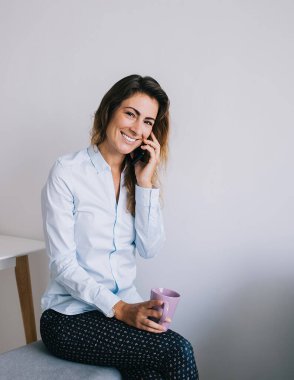 Çekici, genç esmer, gülümseyen ve oturan bacakları çaprazlanmış, elinde kahve fincanıyla büroda beyaz arka planda kameraya bakarken.