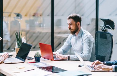 Aktif odaklı erkek ve bayan meslektaşlardan oluşan bir ekip büyük masada oturuyor ve ofiste çalışma sorunlarını tartışırken dizüstü bilgisayarlarda yazı yazıyor.
