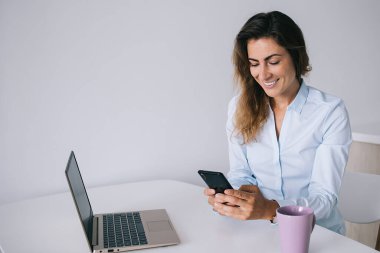 Gömlekli genç esmer, beyaz masada oturuyor, elinde fincan ve laptopla ofisteki akıllı telefondan mesaj atarken gülümsüyor.