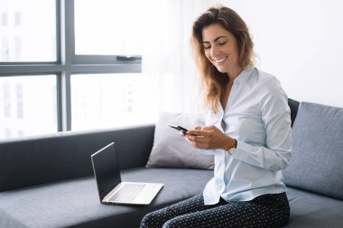 Gömlekli ve pantolonlu genç iş kadını telefonda sohbet ediyor. Oturma odasındaki bilgisayarın yanında oturmuş, gülümsüyor.