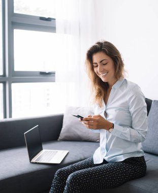 Gömlekli ve pantolonlu genç iş kadını telefonda sohbet ediyor. Oturma odasındaki bilgisayarın yanında oturmuş, gülümsüyor.