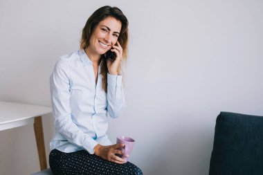 Çekici, genç esmer, gülümseyen ve oturan bacakları çaprazlanmış, elinde kahve fincanıyla büroda beyaz arka planda kameraya bakarken.