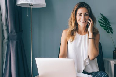 Beyaz kolsuz bluz giyen, yuvarlak masada oturan, akıllı telefonuyla iletişim kuran evde dizüstü bilgisayar kullanan ve kameraya bakan büyüleyici, genç esmer kadın.