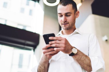 Bulanık arka planda şehir iş merkezinin koridorunda dikilirken akıllı telefonlara bakan modaya uygun beyaz gömlekli mutlu adamdan.