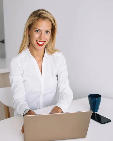 Young female employee sitting at white desk and smiling while working at laptop and looking at camera in office against white wall