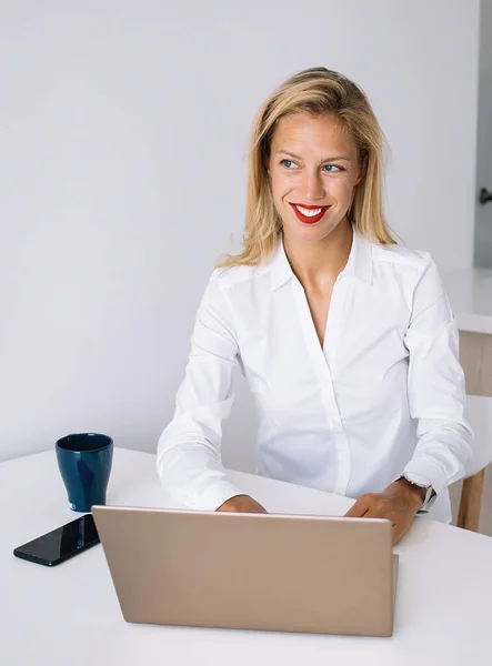 Young blonde in white shirt sitting at desk and smiling while working at laptop and looking away in office against white wall