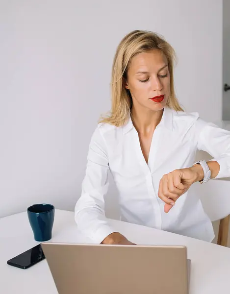 Concentrated young woman sitting at table with laptop and cup while checking time on wristwatch in office against white wall