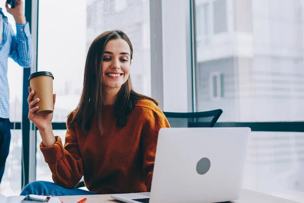 Selective focus on smiling female employee sitting at desktop with cup of coffee satisfied with job in office, happy woman reading good news via laptop while male colleague talking on smartphone