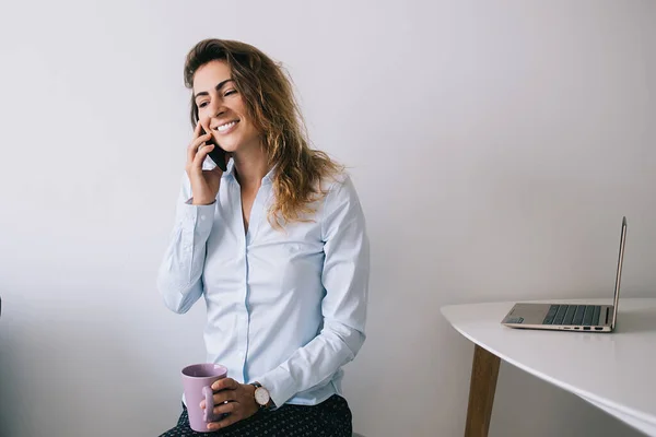 Young pretty female employee smiling and sitting with coffee mug while looking away and talking on phone in office against white background