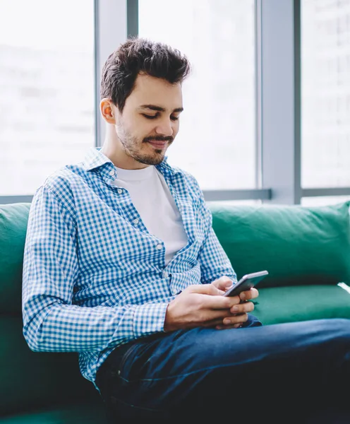 Positive hipster guy dressed in casual wear writing notes in organiser application on cellular phone during resting on comfortable couch, successful male blogger searching information online