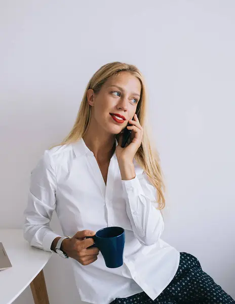 stock image Delighted thoughtful young blonde woman leaning on white table with blue cup in blouse speaking on smartphone using laptop and looking away on white background