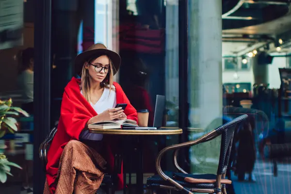 Şapkalı, gözlüklü, renkli kırmızı battaniyeli akıllı telefonlu bir kadın sokak kafesinde açık bir kitap ve dizüstü bilgisayarla otururken akıllı telefonuyla mesajlaşıyor.