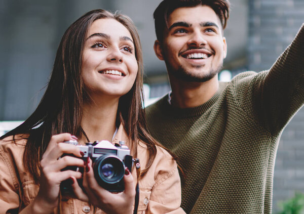Cheerful couple in love spending time together on date using vintage camera for making photos, happy handsome man pointing on smith showing to his girlfriend while teaching photography outdoors