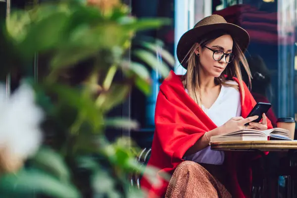 Attentive woman in hat and glasses covered with colourful red blanket messaging smartphone while sitting by table with opened book at street cafe