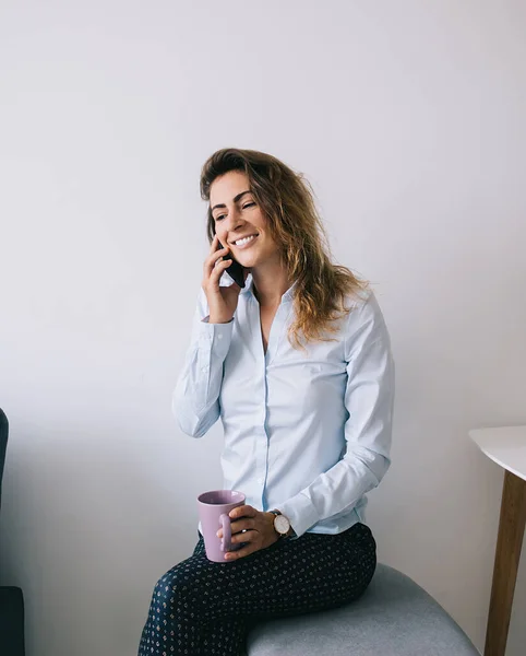 Young pretty female employee smiling and sitting with coffee mug while looking away and talking on phone in office against white background