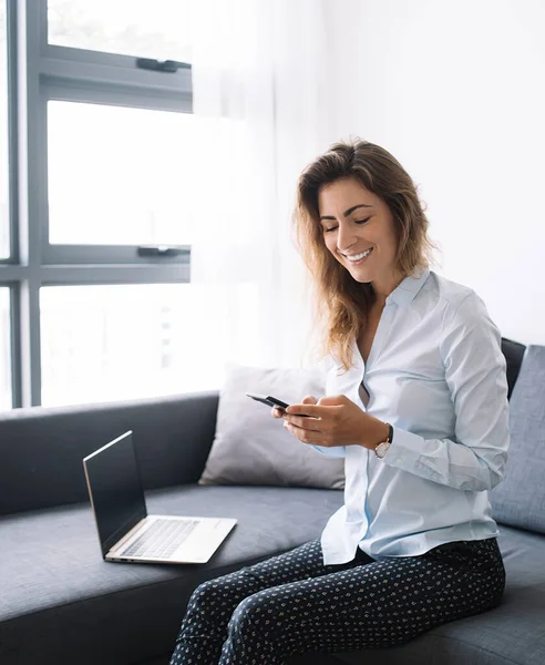 Gömlekli ve pantolonlu genç iş kadını telefonda sohbet ediyor. Oturma odasındaki bilgisayarın yanında oturmuş, gülümsüyor.