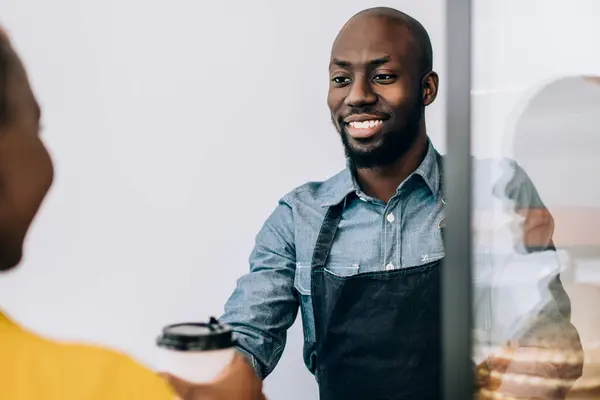 Smiling African American shop assistant in black apron standing at counter and giving disposable cup of coffee to crop customer in pastry shop