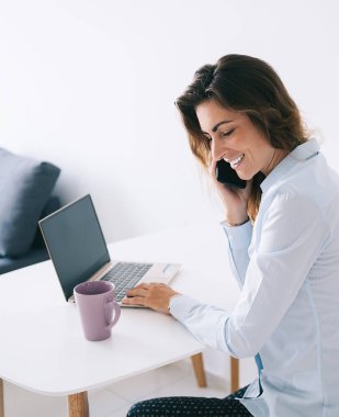 Bardağın ve dizüstü bilgisayarın yanında oturan gülümseyen genç esmerin yan görüntüsü beyaz masada telefonla konuşurken boş bir alan.