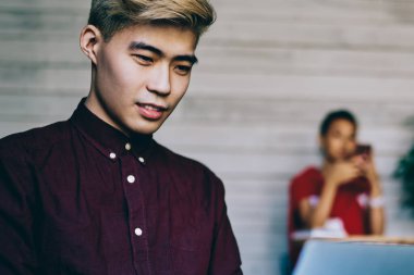 Odaklanmış Asyalı erkek, sarı saçlı, eflatun tişörtlü dizüstü bilgisayarda dizlerinin üzerinde yaratıcı çalışma alanında oturan ve arka planda meslektaşı olan.