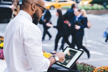 Beyaz tişörtlü, siyah pantolonlu, klavyeli, çiçekli yatağın yanındaki bankta gezen Afro-Amerikan kel adamın yan görüntüsü.