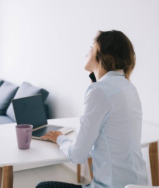 Beyaz masada otururken akıllı telefondan konuşan kupa ve dizüstü bilgisayarın yanında oturan genç kadının yan görüntüsü