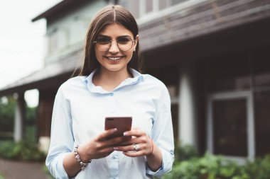 Gözlüklü ve açık renk tişörtlü mutlu bir kadın elinde telefonla kameraya gülümsüyor.