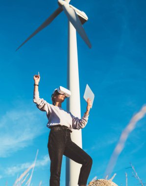 VR gözlük takan genç bir bayan imarcı güneşli bir günde yel değirmeninin yanında dikilirken dokunmatik program ve stil kullanarak sanal siber uzay dizayn ediyor.