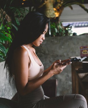 Sportif giyinen kendine güvenen genç bir kadının cep telefonuyla mesaj göndermesi ve gün ışığında rahat bir kafede kahve içmesi yan görünüşü.