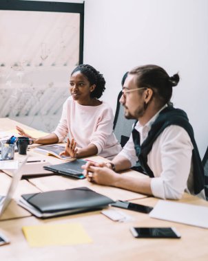Resmi kıyafetlerle yüksek açılı Afrikalı Amerikalı çalışanlar, toplantı sırasında kağıt ve cihazlarla tahta masada otururken çok ırklı meslektaşları için görüşlerini açıklıyorlar.