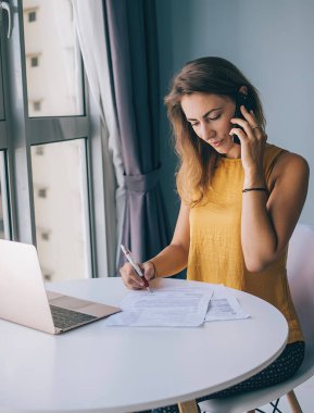 Sarı kolsuz bluzuyla beyaz yuvarlak masada oturan ve evinde akıllı telefondan konuşan dikkati dağılmış genç bir kadın.