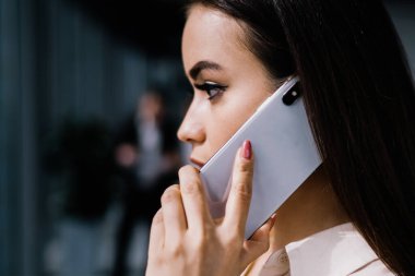 Resmi giyinmiş, modern akıllı telefondan konuşan, iş yerinde dikilip başka yöne bakan çekici, ciddi bir kadının yan görüntüsü.