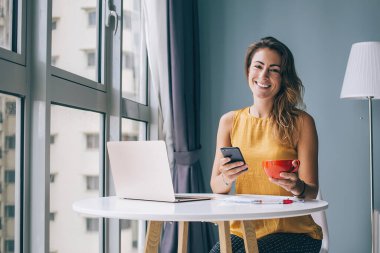 Tasasız, modern, güler yüzlü kadın evinde dizüstü bilgisayarla yuvarlak masada otururken akıllı telefonuyla interneti karıştırıyor.