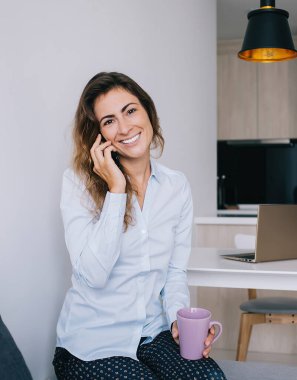 Kahverengi saçlı, bardakta içecekle oturan ve ofisteki kameraya bakıp gülümseyen çekici genç bayan.