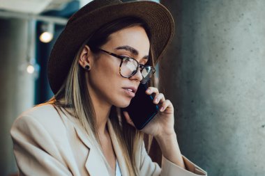 Gözlüklü ve şapkalı zarif yetişkin bir kadının ekrana odaklanırken telefon görüşmesi yapması ve kafenin penceresinin yanındaki bilgisayarı kullanması.