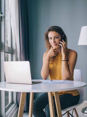 Sarı kolsuz bluzuyla beyaz yuvarlak masada oturan, evinde kameraya bakarak akıllı telefondan iletişim kuran, memnun genç bir kadın.