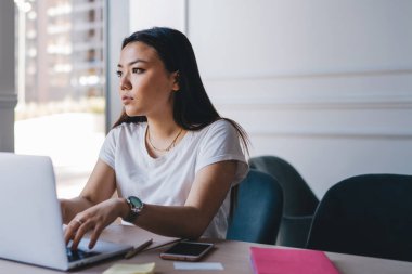Genç, zeki, beyaz tişörtlü Asyalı bir kadın laptopla çalışıyor. Ofisinde akıllı telefon defterleriyle masada oturuyor.