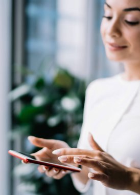 Resmi giysiler içinde genç ve neşeli bir kadın. Bulanık arka planda, pencerenin yanında duran modern akıllı telefona göz atıyor.