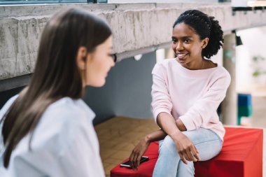 Kırmızı pouf üzerinde bacakları çapraz oturan ve modern dinlenme alanında bayan arkadaşıyla konuşurken gülümseyen Afro-Amerikalı genç bir kadın.