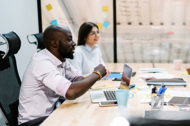 Beyaz gömlekli çok ırklı iş arkadaşları cam duvara karşı projeyi tartışırken dizüstü bilgisayar ve kağıtlarla masada oturuyorlar.