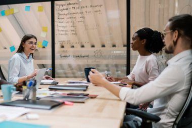 Modern ofiste masa başında oturan iş arkadaşlarıyla ortak bir proje oluştururken gözlüklü, neşeli, pozitif genç bir kadın.
