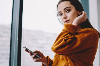 Akıllı telefon cihazı kullanmak için boş zamanlarında kameraya bakarak rahat kapanış giyinmiş ciddi bin yıllık hipster kız yarım uzunlukta portre, cep telefonu ile çekici kadın kapalı poz