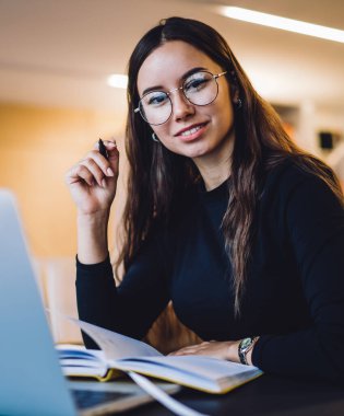 Moda gözlüklü akıllı kadın grafik tasarımcısı serbest çalışma için modern netbook ile birlikte çalışıyor, beyaz kadın blogcunun kameraya gülümseyen portresi