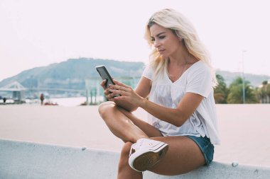 Gündüz kıyafetleri içinde, arkadaşlarıyla mesajlaşarak güneşli bir günde şehir sokaklarında çitlerde oturan ciddi bir genç bayan.