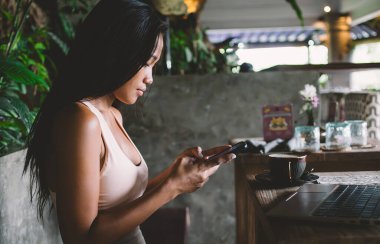 Sportif giyinen kendine güvenen genç bir kadının cep telefonuyla mesaj göndermesi ve gün ışığında rahat bir kafede kahve içmesi yan görünüşü.