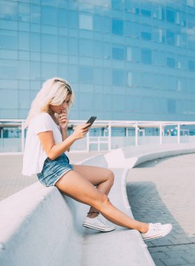 Düşünceli bir kadın, gündelik giysiler içinde telefonla konuşuyor, arkadaşlarıyla iletişim kuruyor, düşünceleriyle ağızlarına dokunuyor, sokakta çitlerde oturuyor.