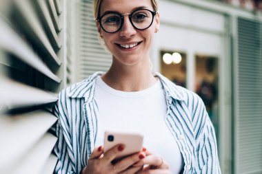 Neşeli sarışın kadın günlük kıyafetler giyiyor ve moda gözlükler takıyor. Gülümsüyor ve kameraya bakıyor. Duvara yaslanıyor ve akıllı telefonlara bakıyor.